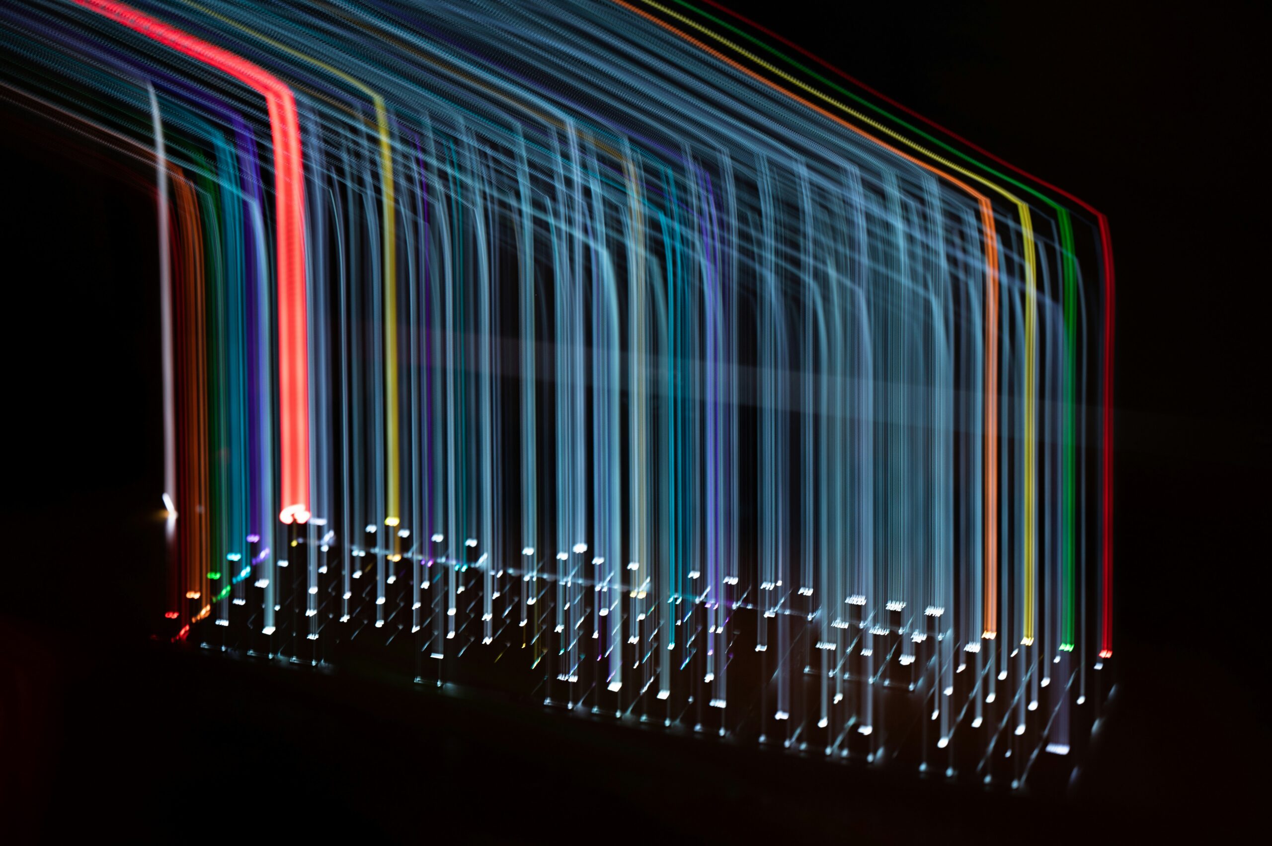Abstract long-exposure image of neon rainbow light trails forming curved vertical lines against a dark background, resembling a light sculpture.